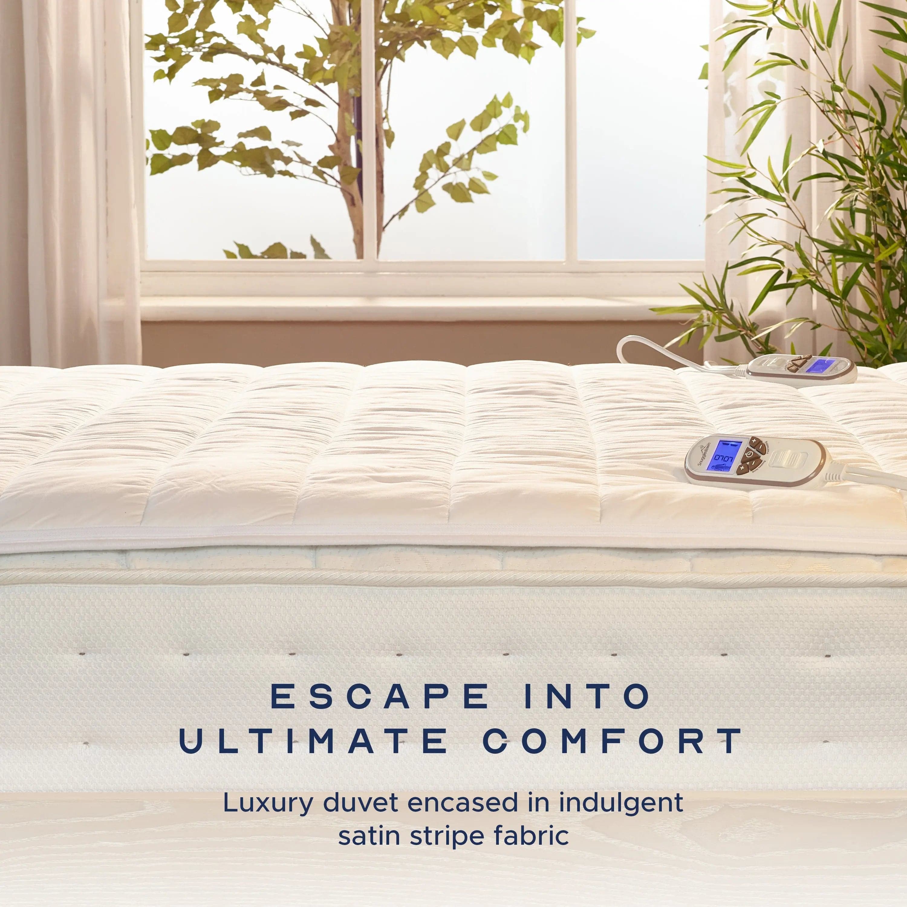 Supreme heated underblanket laid over a bed beside a sunlit window, with two digital controllers resting on the quilted surface.